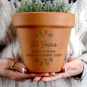 Peut inclure: Pot de fleur en terre cuite avec l'inscription "35 Years", les noms "LINDA & PETER" et la date "23RD NOVEMBER 2026". Le pot contient des plantes vertes et des fleurs violettes. Un cadeau décoratif.
