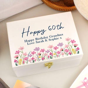 May include: A white wooden box with a gold clasp, adorned with a floral border of pink, purple, and yellow flowers. The box is inscribed with the words "Happy 60th" and a birthday message.