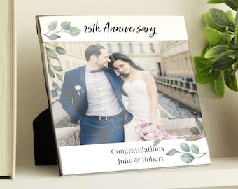 May include: A silver photo frame with a white background and a photo of a couple in front of a building. The frame has a green leafy design and the text "25th Anniversary" and "Congratulations Julie & Robert".