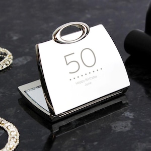 Pode incluir: Um espelho compacto prateado com uma alça, gravado com o número "50" e as palavras "Happy Birthday June". O espelho está aberto, revelando uma superfície refletora. O compacto está sobre uma superfície escura e refletora.