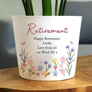 Peut inclure: Pot de fleurs en céramique blanche avec le mot "Retraite" en écriture rose. Le pot est décoré de fleurs à l'aquarelle roses, bleues et jaunes, et le texte "Bonne retraite Linda, de la part de tous au service 3D x".