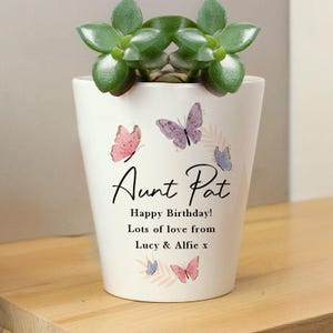 Può includere: Un vaso di ceramica bianco con una pianta succulenta. Il vaso è decorato con farfalle rosa e viola e il testo "Aunt Pat Happy Birthday! Lots of love from Lucy & Alfie x".