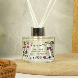 May include: A clear glass diffuser bottle with a silver cap and white reeds. The bottle is decorated with floral designs and text that reads "Happy 100th" and a personalized message. The diffuser sits on a wooden surface.