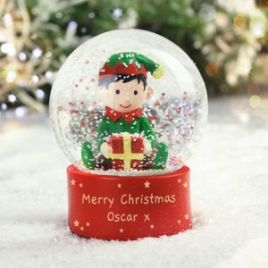 May include: A red and clear glass snow globe with a small elf figurine holding a wrapped gift inside. The base of the snow globe has the text "Merry Christmas Oscar x" written on it.