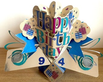 May include: A pop-up birthday card with a blue, silver, and gold design. The card features the words "Happy Birthday" in a colorful font and the numbers "94" on the front.