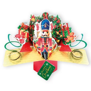 Op de afbeelding: Een pop-up kerstkaart met een gedetailleerde notenkrakersoldaat in een rood en blauw uniform. De kaart bevat kerstbomen, cadeaus en een peperkoekmannetje. Een groen label met "Happy Christmas" is bevestigd.