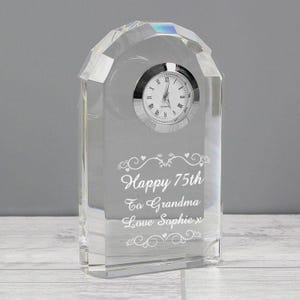 Puede incluir: Un reloj de cristal transparente con esfera plateada y números romanos. El reloj está grabado con las palabras "Happy 75th To Grandma Love Sophie x". El reloj tiene una base rectangular y una parte superior redondeada.
