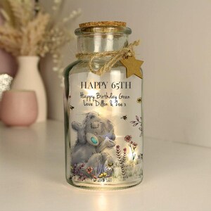 Op de afbeelding: Een glazen pot met een kurkdeksel en een lichtje erin. De pot heeft een bedrukt beeld van een grijze en blauwe teddybeer met bloemen en de tekst "HAPPY 65TH Happy Birthday Gran Love Dillon & Zoe x".