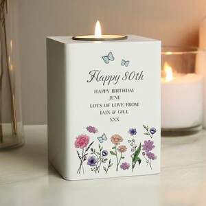 Op de afbeelding: Witte houten kaarsenhouder met een theelichtje erin. De houder heeft een bloemmotief met vlinders en de tekst "Happy 80th Happy Birthday June Lots of Love from Iain & Gill XXX".