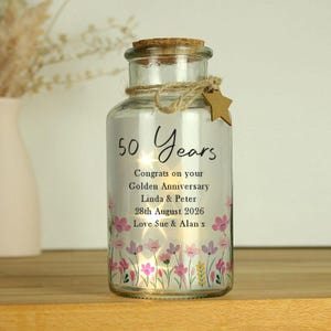 Op de afbeelding: Een heldere glazen pot met een kurk, touw en een stervormig label. De pot is versierd met de tekst "50 Years" en een boodschap voor een gouden jubileum. Roze en gele bloemen staan onderaan.