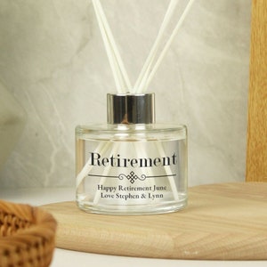 Puede incluir: Una botella de difusor de vidrio transparente con una tapa plateada y texto negro que dice "Retirement" con un diseño decorativo y el mensaje "Happy Retirement June Love Stephen & Lynn".