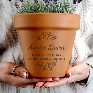 Puede incluir: Una maceta de terracota marrón con el texto "Auntie Laura" y "Happy Birthday Love Emma & Jack X" en escritura negra. La maceta está llena de follaje verde y flores moradas. Hay diseños florales.