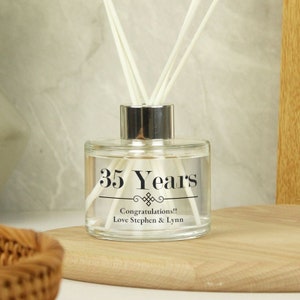 Puede incluir: Una botella de difusor de vidrio transparente con texto negro que dice "35 Years" y "Congratulations!! Love Stephen & Lynn". La botella tiene una tapa plateada y varillas difusoras blancas.