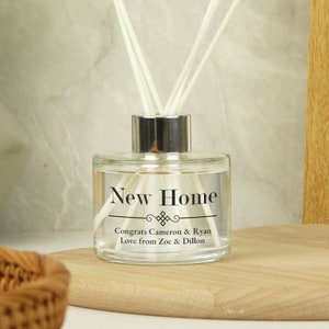 Può includere: Una bottiglia di diffusore in vetro trasparente con un tappo argentato e canne bianche. La bottiglia ha un'etichetta che dice "New Home" con un design decorativo e il testo "Congrats Cameron & Ryan Love from Zoe & Dillon".
