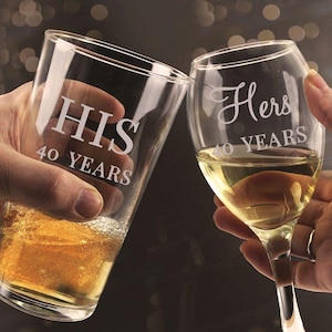 May include: A clear glass pint glass etched with the text HIS 40 YEARS containing a golden liquid and a clear glass wine glass etched with the text Hers 40 YEARS containing a pale yellow liquid, both held up in a toast against a dark background.