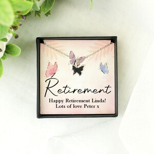 May include: A silver necklace with three butterfly pendants is displayed in a black box. The box features the word "Retirement" and a message. The butterflies are pink, purple, and black.