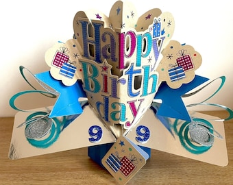 May include: A pop-up card with a heart-shaped design featuring the words "Happy Birthday" in colorful letters. The card is decorated with blue stars, silver glitter, and gift boxes. The card is open and the inside is decorated with a blue and silver swirl design.