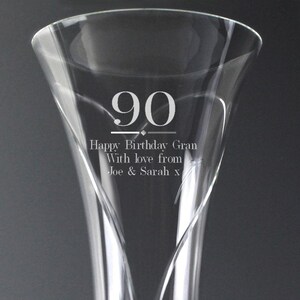 Può includere: Un calice di champagne in vetro trasparente con la parte superiore svasata. Il numero "90" è inciso sul vetro, insieme alle parole "Happy Birthday Gran, With love from Joe & Sarah x". Il bicchiere ha un design a spirale decorativo.
