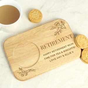 May include: A wooden tea and biscuit serving board with the word "RETIREMENT" engraved. The board has a circular indentation for a teacup and floral designs. Text reads "HAPPY RETIREMENT MUM TIME FOR TEA & BISCUITS! LOVE AMY & ELLIE X".