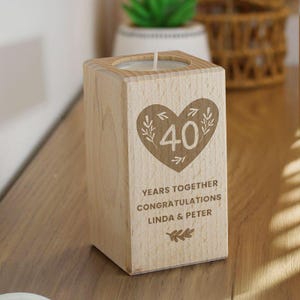 May include: A wooden candle holder engraved with a heart design, the number 40, and the text "YEARS TOGETHER CONGRATULATIONS LINDA & PETER". The candle holder is made of light wood and is square-shaped.