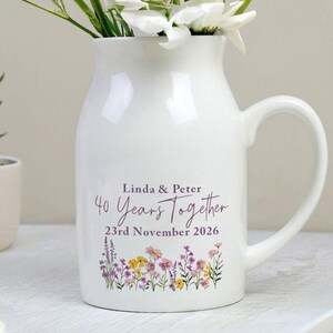 Puede incluir: Un jarrón de cerámica blanco con asa, lleno de flores blancas. El jarrón está decorado con el texto "Linda & Peter 40 Years Together 23rd November 2026" y un diseño floral en morado, amarillo y rosa.