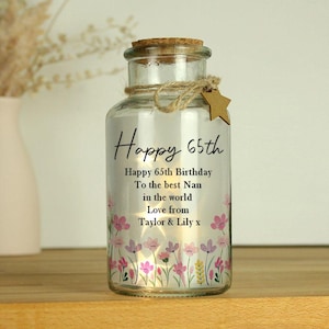 Op de afbeelding: Een heldere glazen pot met een kurk, touw en een stervormig label. De pot is versierd met de tekst "Happy 65th" en een verjaardagsboodschap. Roze en geel bloemmotief aan de onderkant.
