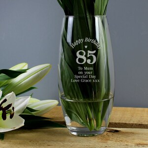 Könnte beinhalten: Eine klare Glasvase mit geschwungener Form, gefüllt mit Wasser und grünen Blättern. Die Vase trägt den Text "Happy Birthday 85" und eine Nachricht für Mama. Weiße Lilienblüten sind links zu sehen.
