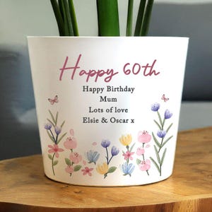 May include: White ceramic plant pot with the text "Happy 60th" in pink script. The pot is decorated with watercolor flowers in pink, blue, and yellow, and the words "Happy Birthday Mum, Lots of love, Elsie & Oscar x".