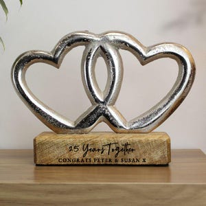 Op de afbeelding: Een zilveren sculptuur van twee in elkaar grijpende harten op een houten voet. De voet is gegraveerd met de tekst "25 Years Together" en "Congrats Peter & Susan X". Een decoratief item voor een jubileum.
