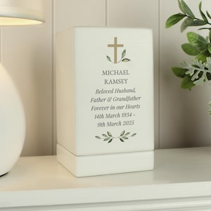 Puede incluir: Caja de madera blanca con una cruz dorada y hojas verdes. La caja tiene el texto "MICHAEL RAMSEY Beloved Husband, Father & Grandfather Forever in our Hearts 14th March 1934 - 9th March 2025" en ella.