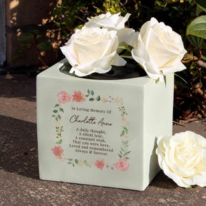 Può includere: Un vaso commemorativo quadrato verde chiaro con rose bianche. Il vaso presenta un motivo floreale e il testo "In Loving Memory Of Charlotte Anne". Il testo include le frasi "A daily thought, A silent tear, A constant wish, That you were here, Loved and remembered Always & forever."