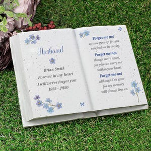 May include: A white, open book-shaped memorial stone with blue floral accents and the words "Husband" and "Forget me not" inscribed. The text includes a name, dates, and a heartfelt message, suitable for remembrance.