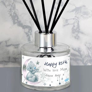 Op de afbeelding: Glazen diffuserfles met zwarte stokjes. Het etiket luidt "Happy 85th With love Mum From Amy". Een cartoonbeer zit op het etiket, op een halve maan. De diffuser heeft een zilveren dop.