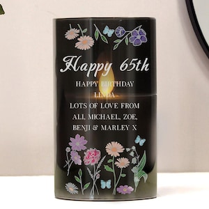 Op de afbeelding: Een zwarte kaars versierd met bloemen en vlinders. De kaars heeft de tekst "Happy 65th Happy Birthday Linda Lots of Love From Ali, Michael, Zoe, Benji & Marley X". De kaars heeft een warm, flikkerend licht.