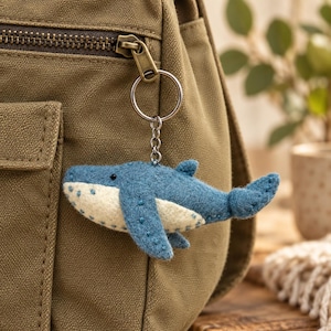 May include: A blue and cream-colored felt whale keychain attached to a silver-toned metal ring and chain. The whale features visible stitching along its fins and body, with a small black bead for an eye, hanging from a brown canvas bag.