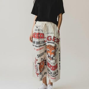 Puede incluir: Una camiseta negra de manga corta combinada con pantalones anchos de color blanco roto con cintura de cordón. Los pantalones están decorados con grandes tipografías de estilo vintage en rojo y negro e ilustraciones gráficas de cabezas de tigre.