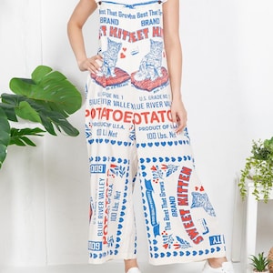 May include: Cream-colored jumpsuit with a vintage potato sack design. The jumpsuit features a print of two kittens, text that reads "Sweet Kitten Brand", and "Blue River Valley" branding. The jumpsuit is paired with a white t-shirt and white sneakers.