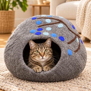 May include: A grey felted cat cave with a dome shape and a circular opening. The exterior features a brown branch design with circular blue and light blue felt accents. A brown tabby cat is resting inside the cave, looking toward the front.