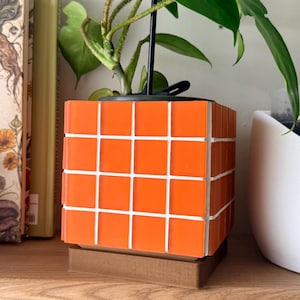 May include: A square plant pot featuring a grid of bright orange tiles separated by white grout lines. The pot sits on a brown square base and contains a green plant with a black support stake extending from the center of the soil.