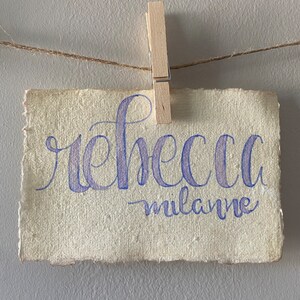 May include: A rectangular piece of off-white textured paper with deckled edges, featuring the name "rebecca milanne" written in purple cursive calligraphy. The paper is secured to a thin brown twine string by a light-colored wooden clothespin.