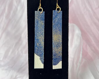 May include: A pair of long, rectangular drop earrings featuring a blue and white marbled pattern with gold glitter accents. The earrings are suspended from gold-toned metal hooks and have a smooth, flat surface with a distinct color-blocked design.