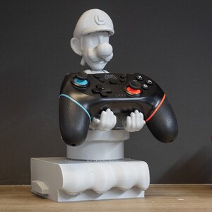 Puede incluir: Busto gris impreso en 3D de un personaje de dibujos animados que sostiene un mando de videojuegos negro. El mando tiene detalles en azul y rojo. El busto está sobre una base blanca.