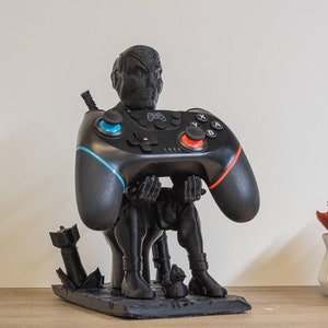 May include: A black gaming controller holder featuring a stylized figure holding a black controller with blue and red accents. The figure is black and sits on a rectangular base with a bomb and other details.