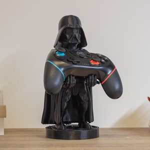 May include: A black Darth Vader figurine holding a black video game controller with blue and red accents. The figurine is on a round black base. The controller has multiple buttons and directional pads. The figurine is on a wooden surface.