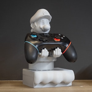 Puede incluir: Una estatua gris impresa en 3D de un personaje de dibujos animados que sostiene un mando de videojuegos negro y rojo. El mando tiene detalles en azul y naranja. La estatua está sobre una base cuadrada.