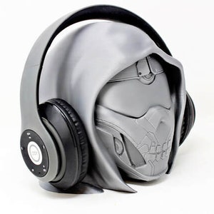 May include: A grey and black headphone set with a distinctive design. The headphones are integrated into a sculpted head, featuring a hood and a detailed mask. The design is predominantly grey with black earcups and controls.