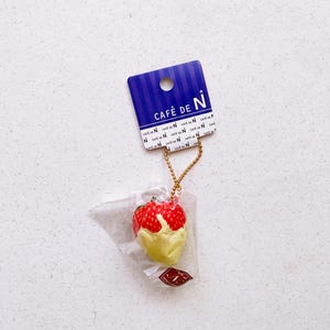 May include: A strawberry-shaped charm with red and white details, dipped in yellow cream, attached to a gold chain. The charm is packaged in clear plastic and hangs from a blue tag with "CAFÉ DE N" text.