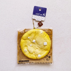 May include: A yellow, round, squishy toy resembling a melon pan, with white square accents. It is packaged in a clear bag with newspaper-style print. A blue tag with "CAFÉ DE N" is attached.