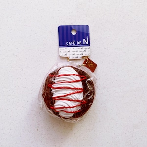 May include: A chocolate-colored pastry with white frosting and red drizzle, packaged in clear plastic. The tag reads "Café de N" in white text on a blue background.