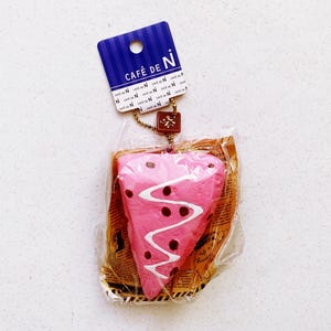 May include: A pink, heart-shaped cake slice squishy toy with white icing and brown dots, packaged in clear plastic. A blue tag with "CAFÉ de N" is attached with a chain.